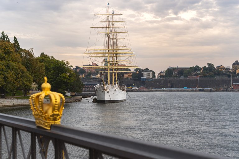 The large, white ship "af Chapman" docked by the green, small island of Skeppsholmen. The photo is taken from the grey steel bridge leading to the island, and the railing is adorned with a golden crown.