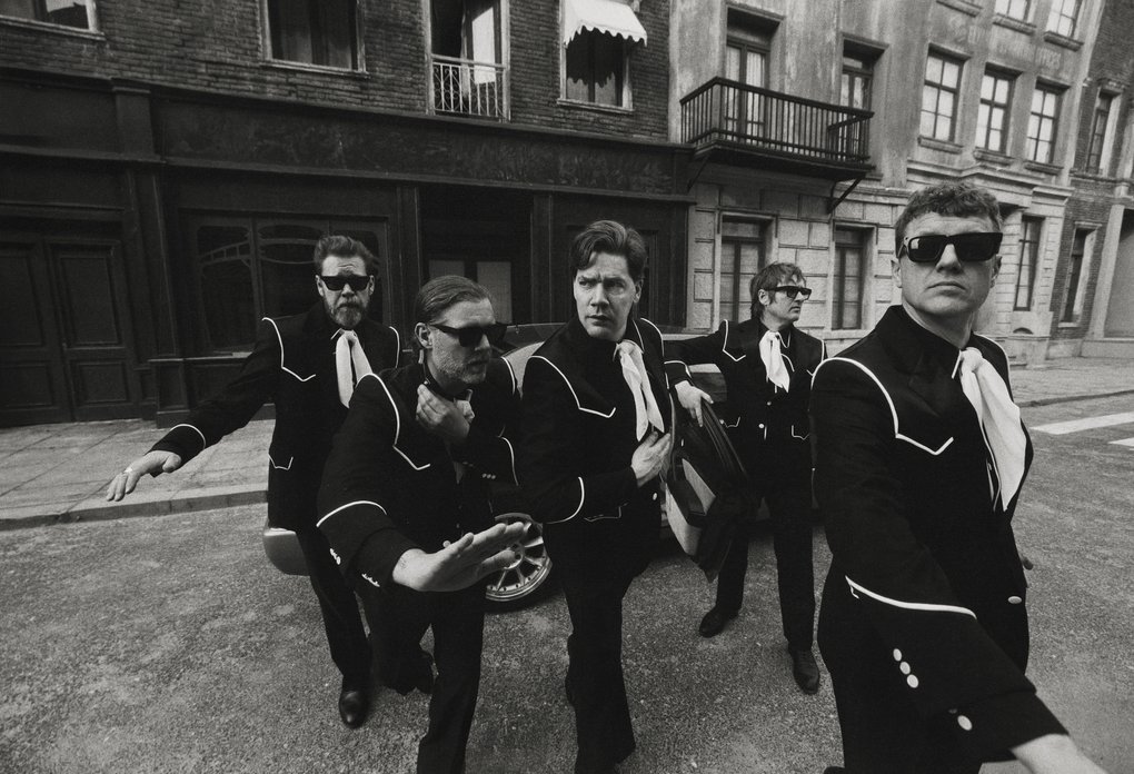 Five band members in matching black suits with white trim and ties, wearing sunglasses, striking dynamic poses on a city street; black-and-white photo.