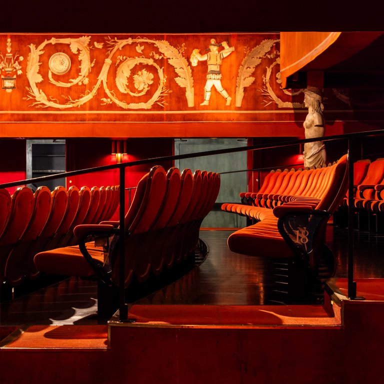 Interior of Skandiabiografen with red cinema chairs seen from the side. In the background you can see a statue and detailed art on the walls.