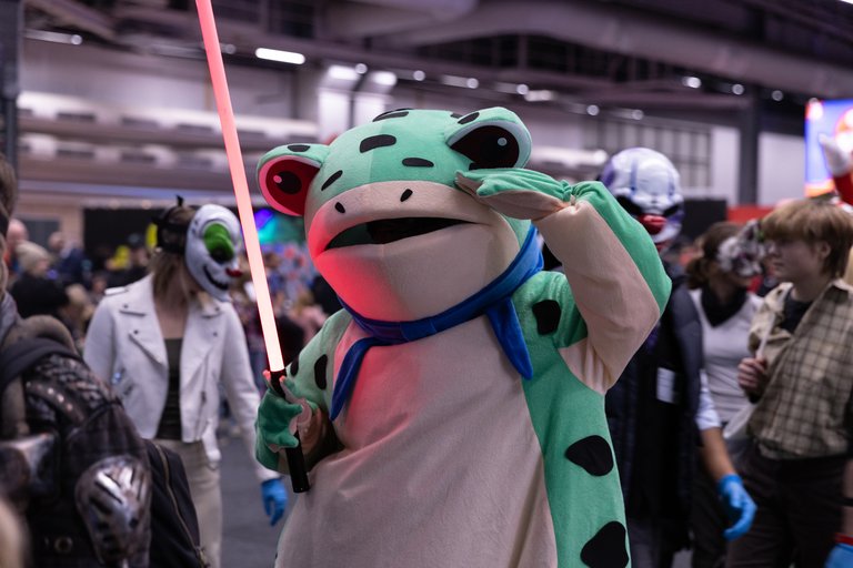 A person is dressed in a frog costume, holding a glowing light saber prop, at Comic Con in Stockholm.