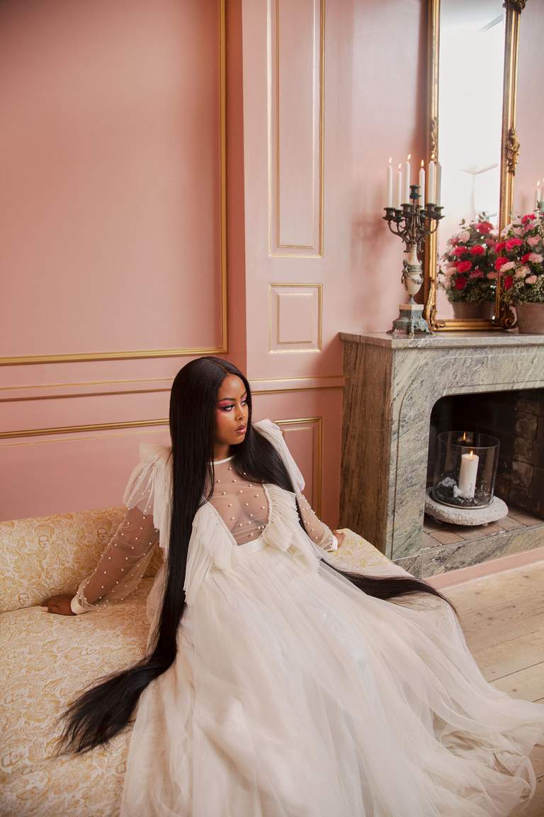 The artist Cherrie in a long white dress sitting on a sofa.