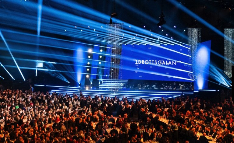 Seated gala dinner guests in front of a stage, and a screen with the word "Idrottsgalan" in white text on a blue background.