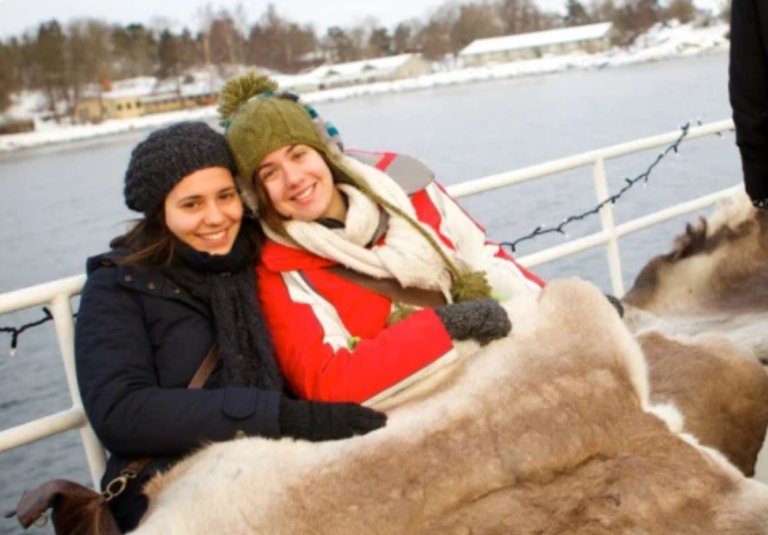 Two people, wearing winter jackets smiling at the viewer whilst seated on a boat. In the background, a snowy Stockholm shoreline can be seen over the water.