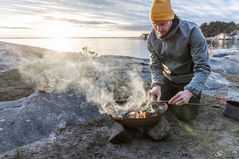 Man in a yellow beanie lighting a fire in a fire bowl by the shore at sunset