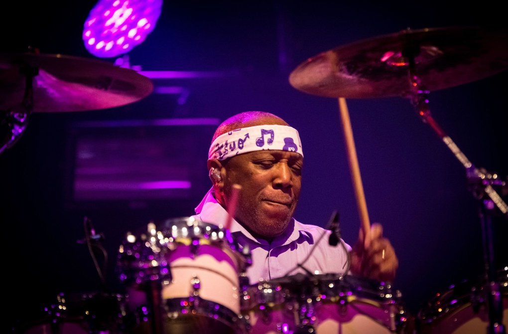 Trummisen Billy Cobham, spelar intensivt bakom ett trumset på en scen upplyst av lila och rosa scenljus. Han bär en vit pannband med noter och håller trumpinnar i rörelse. Bilden förmedlar energi, rytm och livekänsla.
