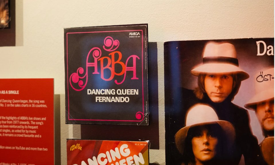 The image shows a vinyl record and a CD case for ABBA's song Dancing Queen along with another record cover featuring a man in a hat. The records are displayed on a shelf with informational text beside them.