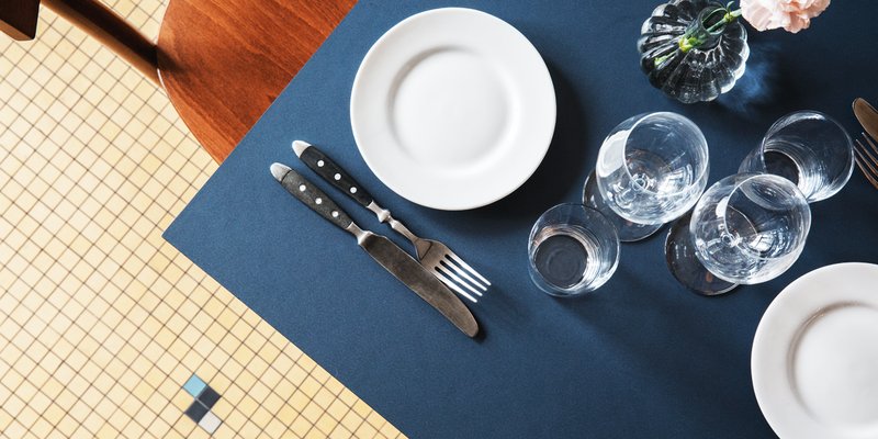 A table set for one, glasses and cutlery against an indigo blue table. A pale pink rose in a vase decorates the table at Bar Agrikultur.