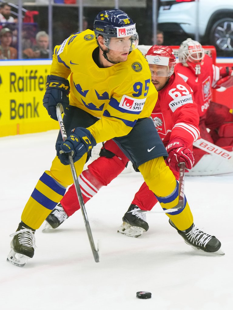 Hockey players during a game. A player dressed in Swedish colors is driving the puck, with an Austrian player coming up behind him.