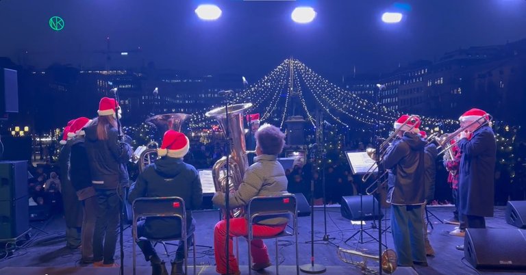 A brass ensemble performing on stage viewed from behind, with light decorations and winter-clad audiences in the background.