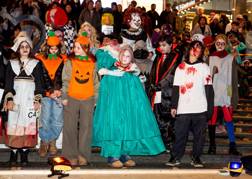 A group is posing in various Halloween costumes on a stage with an audience in the background. The costumes include jack-o'-lantern themes, bloodstains, theatrical outfits, and horror masks.