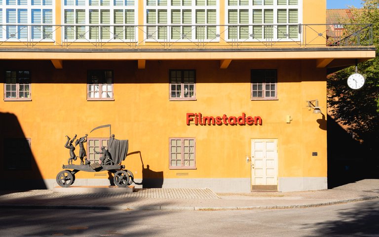 A statue stands in front of an orange building. The statue depicts Ingmar Bergman in front of a circus carrige.