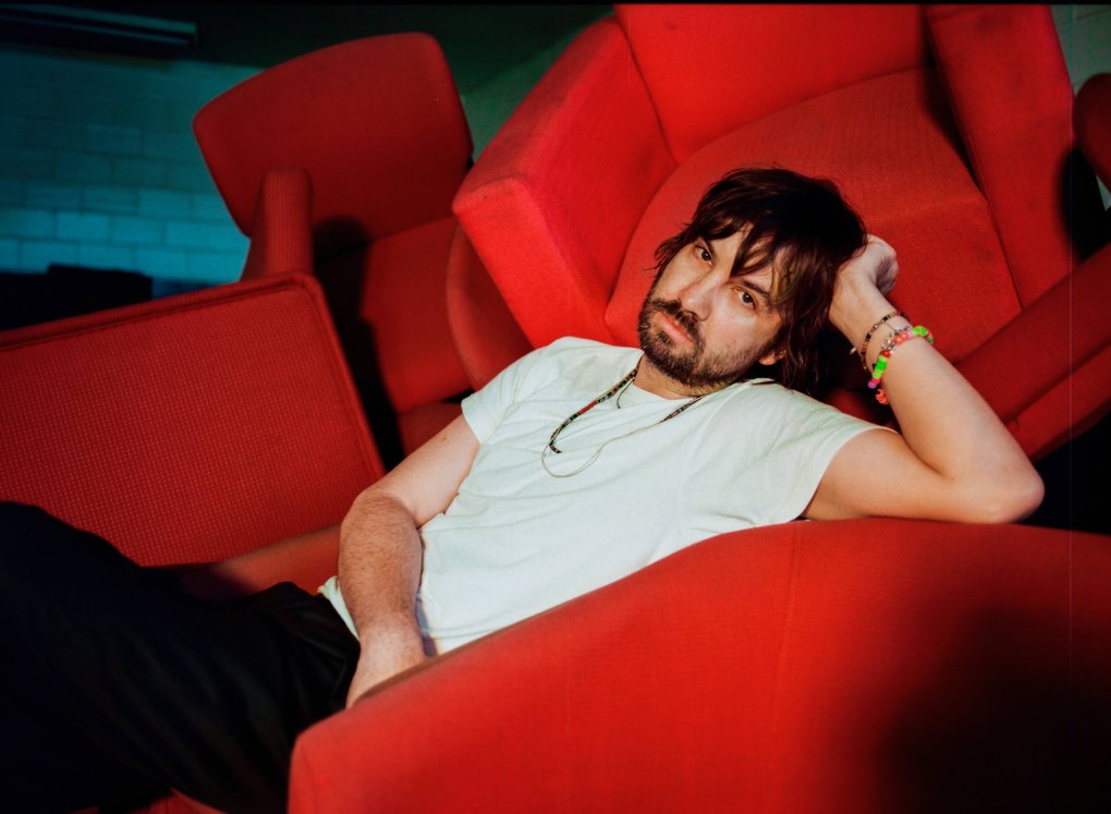 A man in a white t-shirt and colorful bracelets, posing in a sea of red furniture.