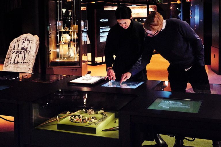 Two people standing in the middle of the exhibition about Vikings, peering over an illuminated glass cabinet