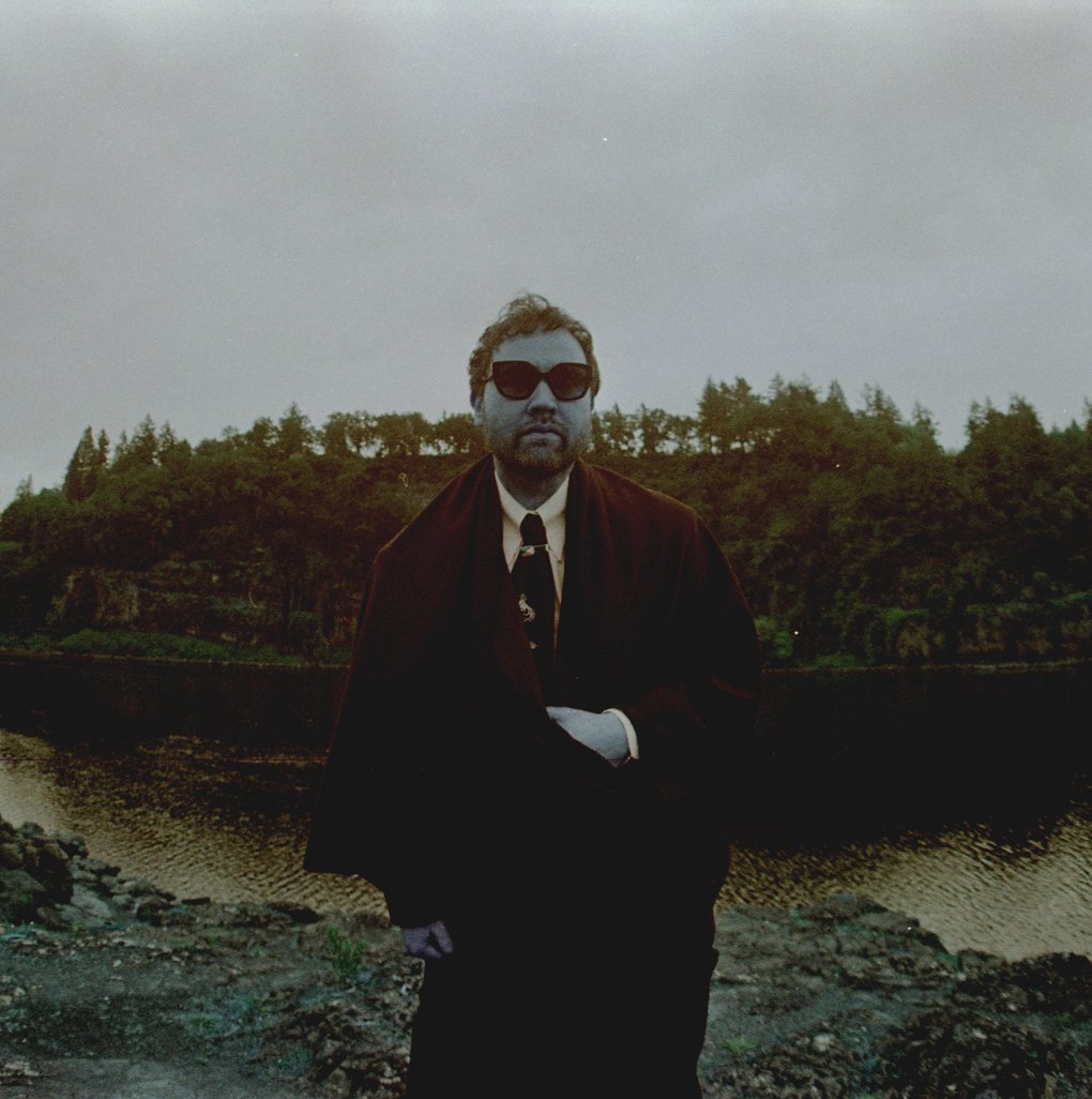 A man wearing a suit and sunglasses standing on a rocky surface above a river, with a woodland landscape visible on the other side.