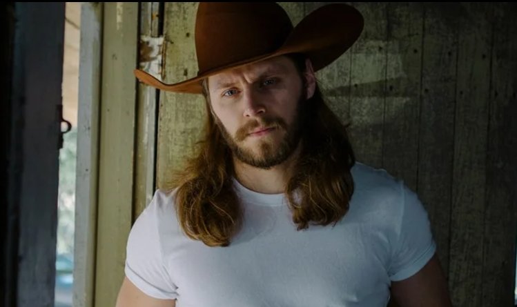 Portrait shot of artist Warren Zeiders, sporting long hair and beard, wearing a white t-shirt and brown cowboy hat.